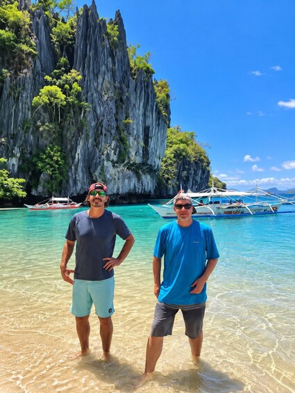 Two people standing in shallow water with a rocky cliff and boats in the background. 5