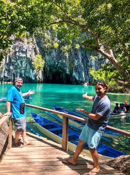 Two people standing on a wooden dock by a turquoise body of water with boats, surrounded by lush greenery and rocky cliffs. 2