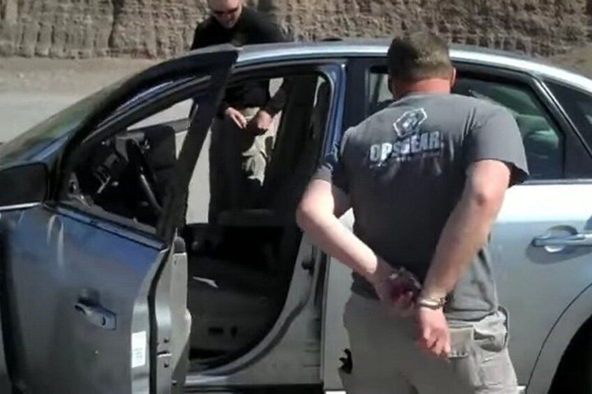 A man in a grey shirt with 'OPSEC' on the back standing next to a car with its door open. 3