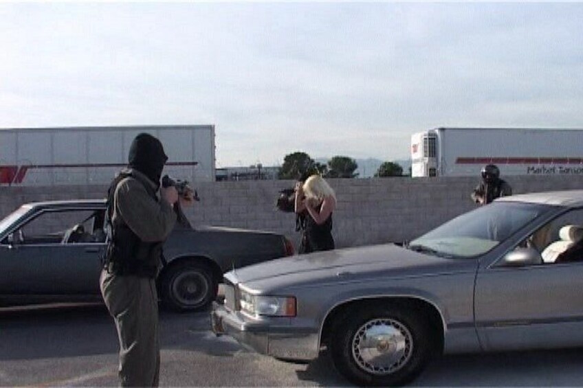 A person in tactical gear aiming a firearm, a parked car, and a building with a sign. 1