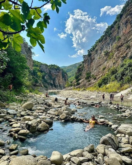 A river with people swimming and sitting on rocks, surrounded by cliffs and greenery. 1