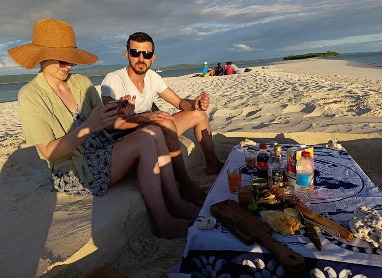 A beach scene with people sitting on the sand, a table with food and drinks, and a body of water in the background. 3