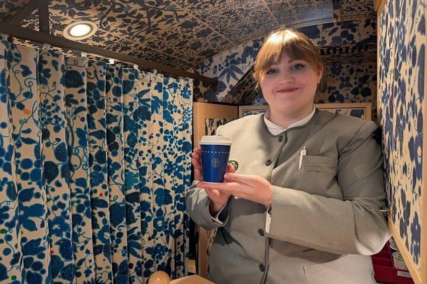 A person holding a blue cup with a lid, standing in a room with patterned wallpaper and a ceiling with a decorative design. 2