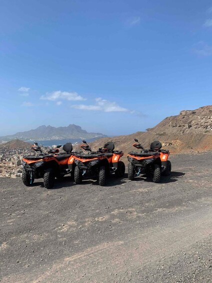 Three orange ATVs parked on a rocky terrain with a cityscape and mountains in the background. 4