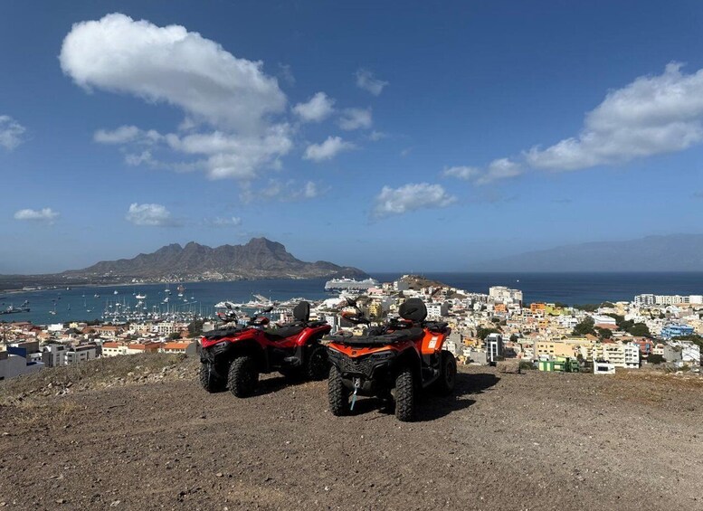 Two red off-road vehicles parked on a rocky hill overlooking a coastal town with buildings and boats in the harbor. 1