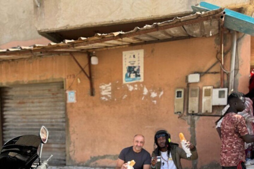 Un groupe de personnes est assis et mange près d’un bâtiment dont le toit est fait de tôles ondulées et dont la porte est fermée par un volet. 2