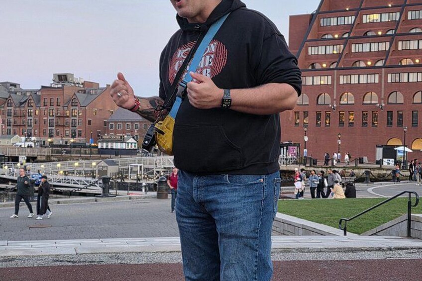 A man standing on a bridge with a camera, buildings, and a waterfront in the background. 4