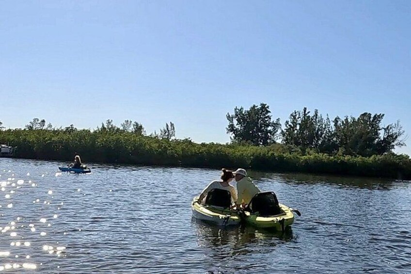 Zwei Personen in einem gelben Kanu auf einem ruhigen Fluss, eine weitere Person in einem blauen Kanu im Hintergrund, umgeben von Grünpflanzen. 5