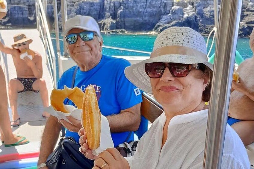 En person holder et stort stykke friturestegt mad, mens en klippefyldt kystlinje og klart, blåt vand ses i baggrunden. 3
