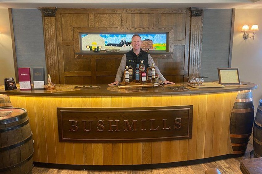 Eine Holztresenbar mit einem Schild mit der Aufschrift 'BUSHMILLS' und einem an der Wand befestigten Fernseher. 2