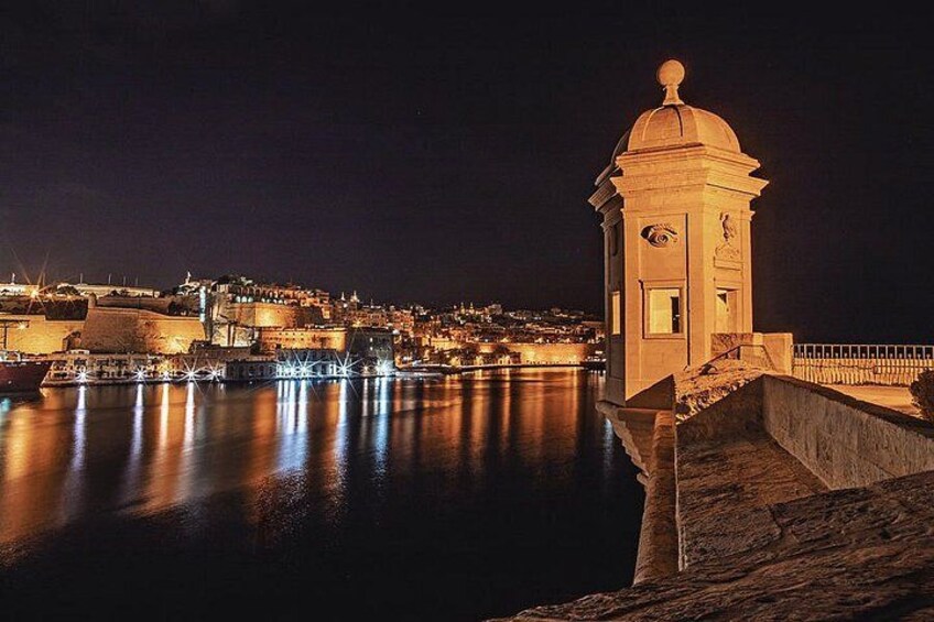 Une scène nocturne d’une ville côtière, avec une tour imposante, des eaux calmes reflétant les lumières et un mur fortifié. 1
