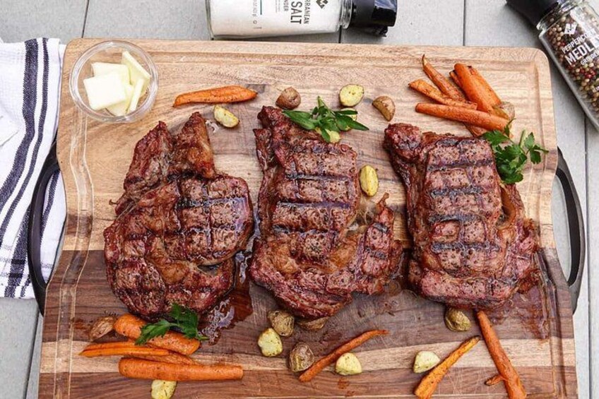 Trois steaks grillés sur une planche à découper en bois, accompagnés de carottes et d’herbes aromatiques. 4