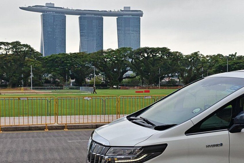 Une voiture blanche portant la mention « HYBRIDE » est garée devant un grand immeuble composé de trois tours, lequel est entouré d’une zone délimitée par une clôture et plantée d’arbres. 1