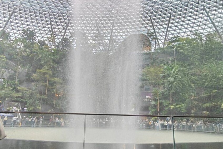 Une fontaine d’eau surmontée d’un toit en verre aux formes géométriques, entourée de verdure. 3