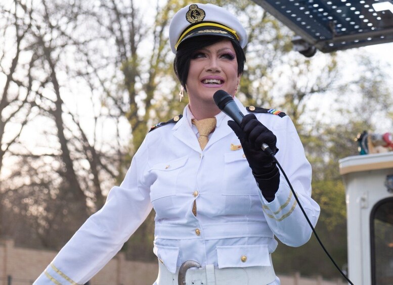 A person in a white uniform with gold buttons and a cap, holding a microphone. 1
