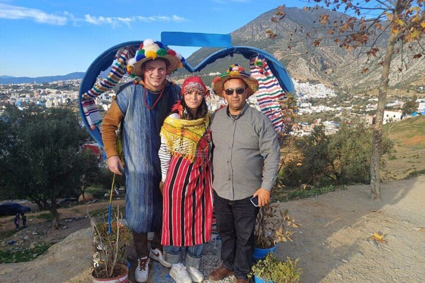 Drei Personen in traditioneller Kleidung vor einem blauen Bogen, im Hintergrund eine gebirgige Landschaft. 3