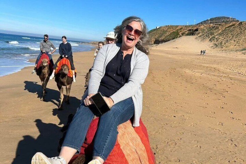 En person sitter på en röd och vit filt på en strand, med hästar och ryttare i bakgrunden. 1