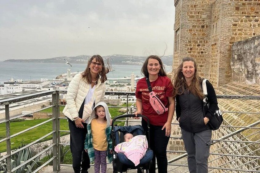 Un grupo de personas de pie en una escalera con vista a un paisaje urbano costero y un edificio histórico de piedra al fondo. 5