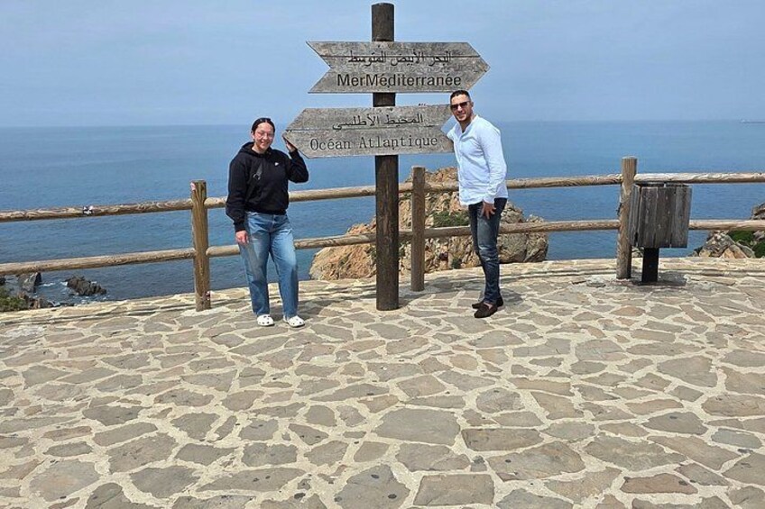 Dos personas de pie frente a un área empedrada de piedra, con una cerca de madera y un poste con varias indicaciones. 2