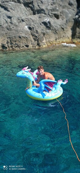 A person on a blue inflatable dragon float in clear blue water with rocky cliffs in the background. 2