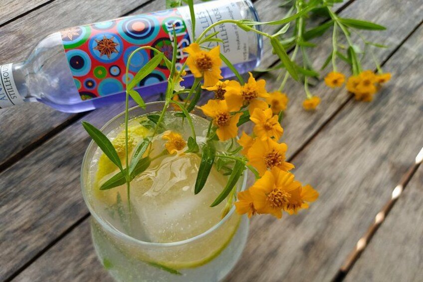 Un vaso de bebida helada con flores amarillas y hojas de menta, una botella de agua Stillhouse, y una mesa de madera. 3