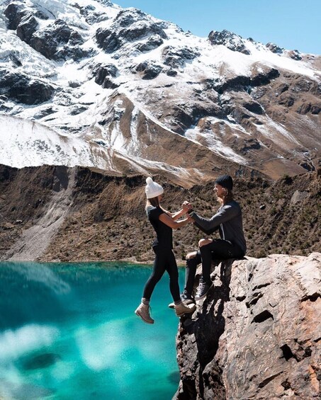 Deux personnes sont assises sur un promontoire rocheux, au bord d’un lac turquoise, avec des montagnes enneigées en arrière-plan. 3