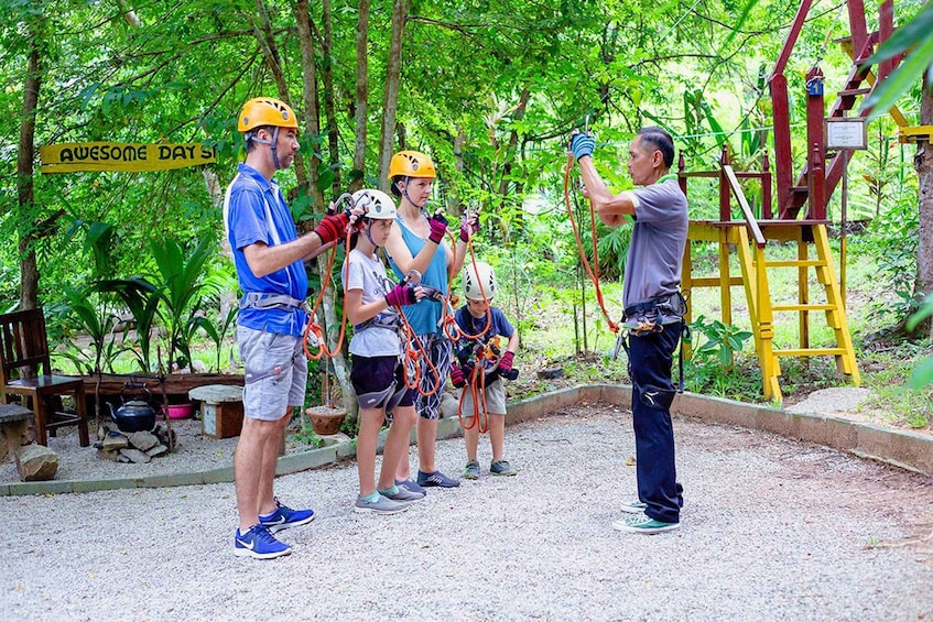 A group of people preparing for a zip-lining activity in a forested area with a sign that reads "AWESOME DAY ST". 2