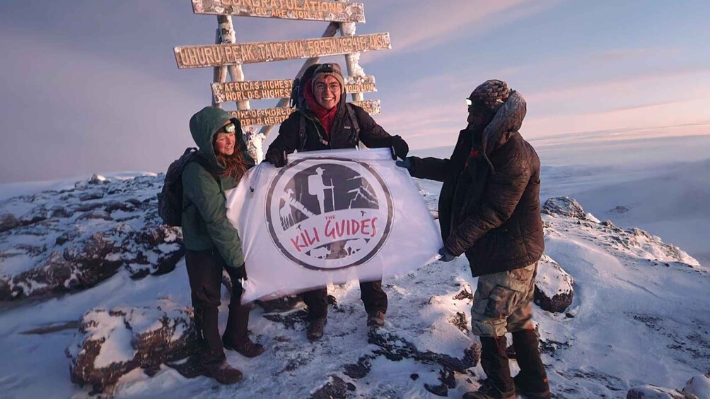 Three people on a snowy mountain holding a banner with a logo. 1