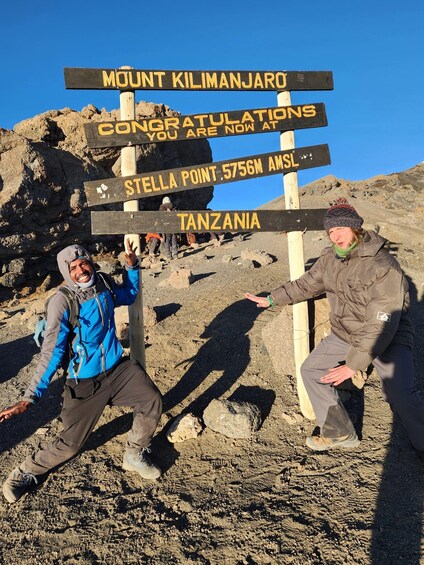 Two people posing in front of a signpost with directions to Mount Kilimanjaro, Stella Point, and Tanzania. 2
