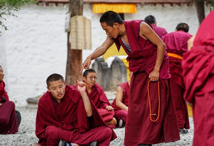 Monks in red robes sitting and standing on a stone surface, with one monk holding a prayer bead. 5