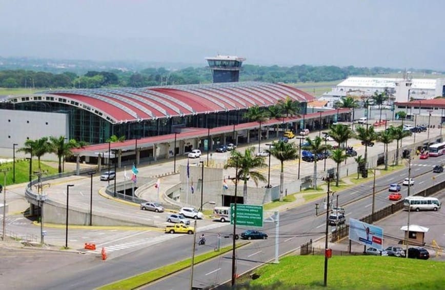 A large airport terminal with a curved roof, surrounded by palm trees and a busy road with vehicles. 2