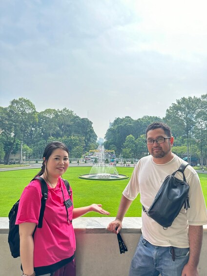 Dos personas junto a una fuente en un parque, con árboles y una carretera al fondo. 4