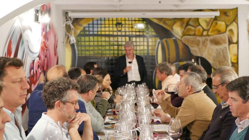 A group of people seated at a long table with wine glasses and plates, in a room with a mural and a stone wall. 5