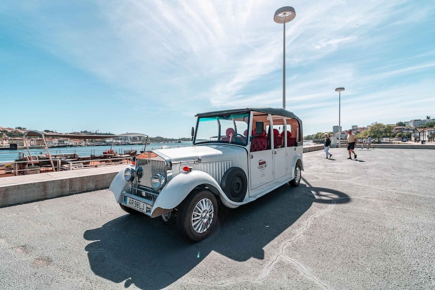 A vintage white car parked near a waterfront with people walking in the background. 3