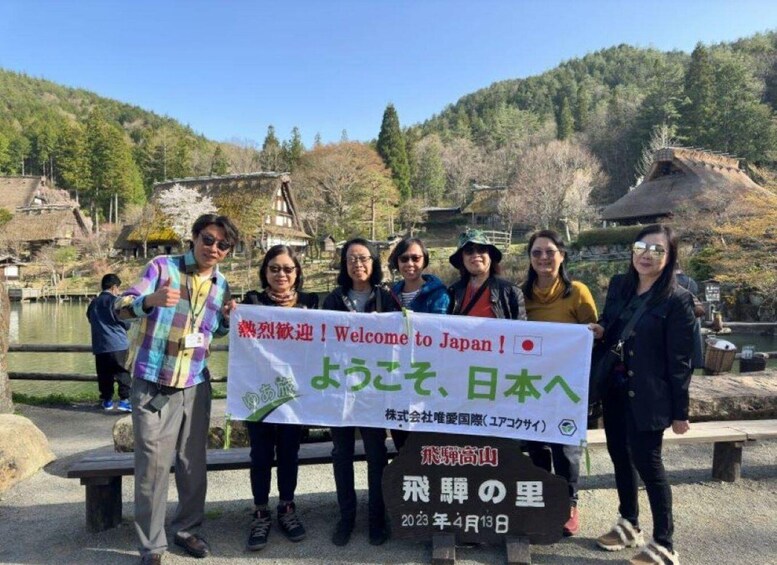 A group of people holding a banner welcoming visitors to Japan, standing in front of a scenic landscape with a pond and traditional buildings. 4