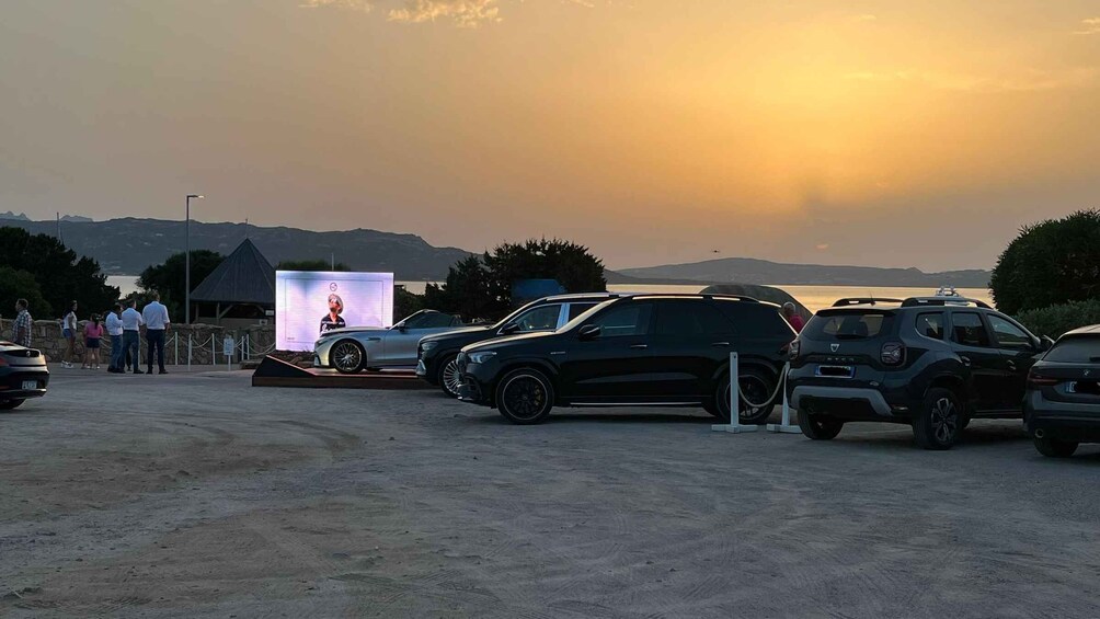 A car show with several vehicles on display, a large screen with an image, and a sunset in the background. 2