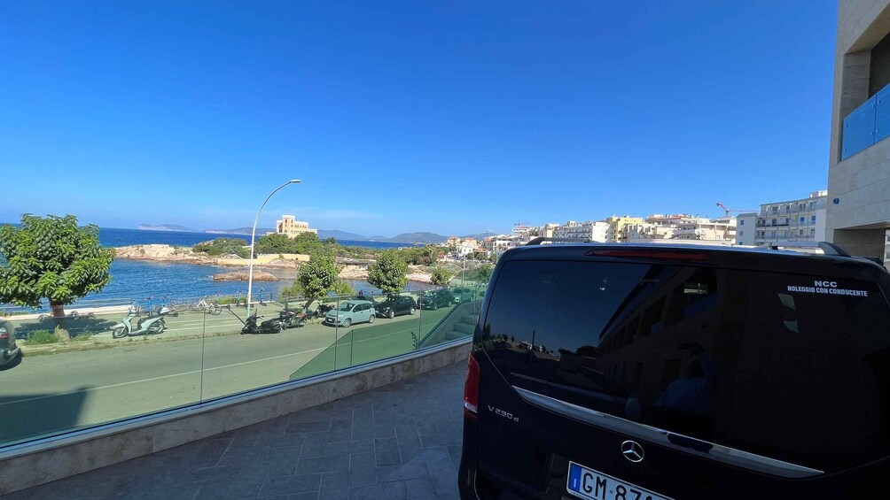 A black van parked on a balcony overlooking a coastal area with a clear blue sky, a tennis court, and buildings in the background. 3