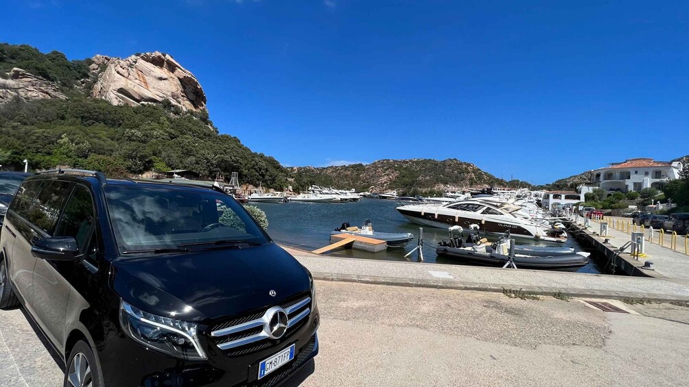 A black car parked by a marina with boats docked in the background. 1