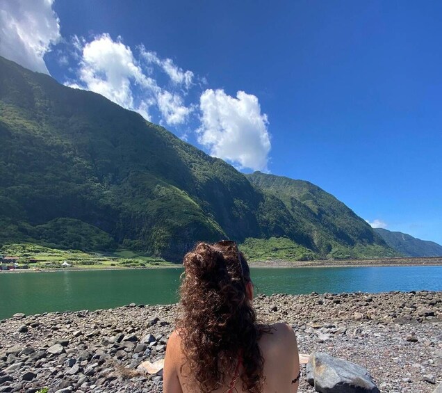 A person sitting on a rocky shore with a mountainous landscape and a body of water in the background. 1