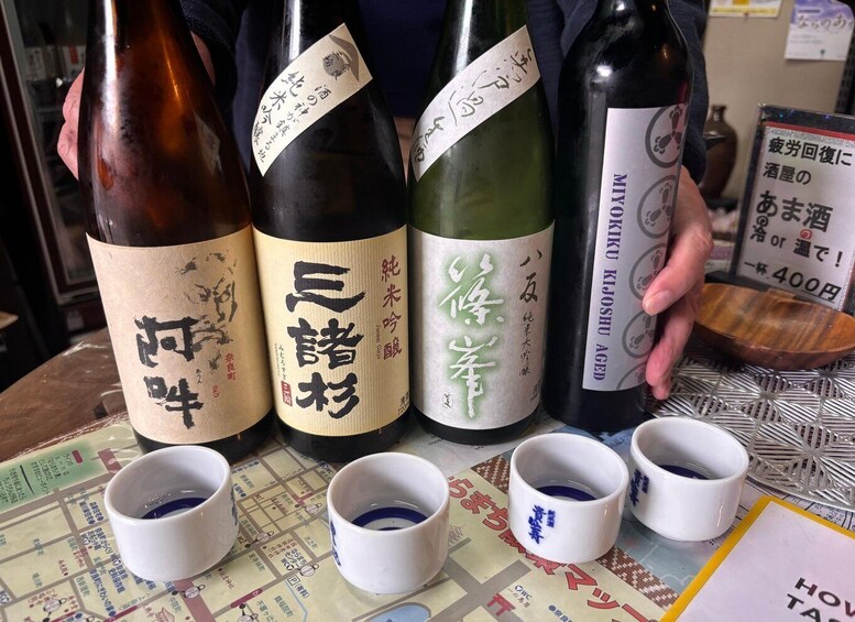 Four bottles of sake with Japanese labels, and five small cups with blue patterns on a table. 1