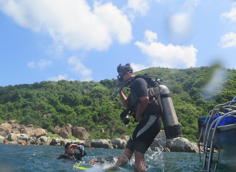 Un sub con bombola d'ossigeno che si prepara ad entrare in acqua, un altro sub già immerso, e una costa rocciosa con una vegetazione rigogliosa sullo sfondo. 2