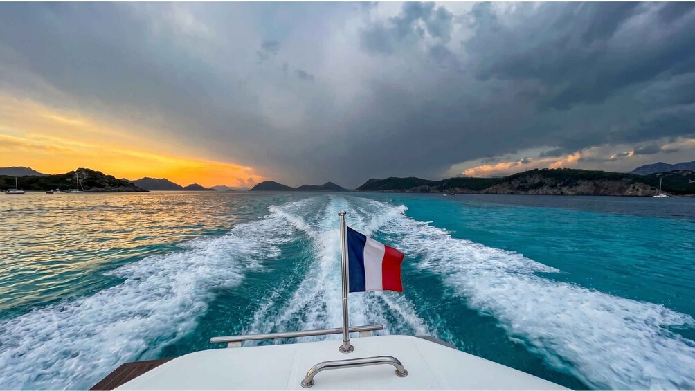 A boat's wake in turquoise waters with a French flag, mountains in the distance, and a cloudy sky. 2
