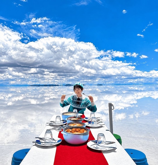 A person sitting at a table with a meal, set against a vast, reflective landscape under a blue sky with clouds. 2