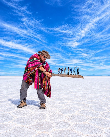 A person in colorful traditional attire holding a long pole, standing in a vast, flat, white landscape under a blue sky with scattered clouds. 1