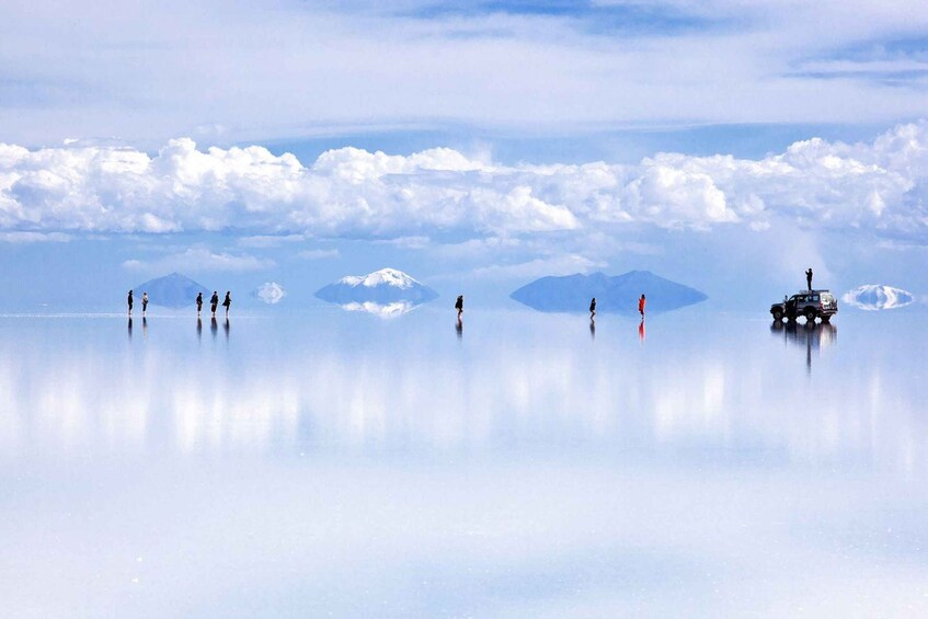 A calm, reflective lake with several people standing on the shore and a vehicle on the water. 1