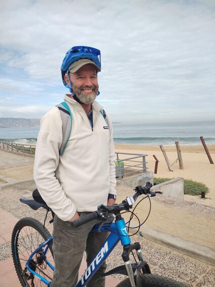 A man standing next to a blue bicycle with a helmet, a beach, and the ocean in the background. 2