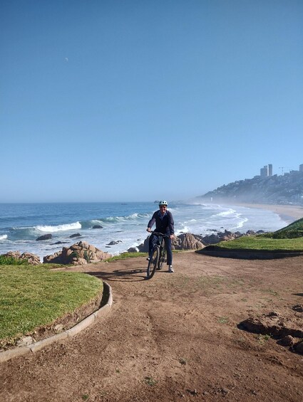 A person riding a bicycle on a dirt path near the ocean, with waves crashing onto the shore and buildings in the background. 4