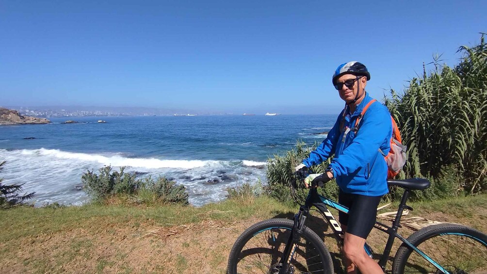 A person standing next to a mountain bike overlooking a coastal view with waves and a distant city skyline. 3