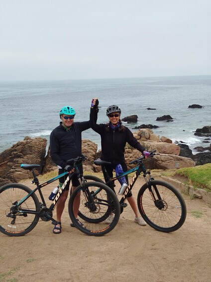 Two people standing on a rocky shore with bicycles, ocean in the background. 4