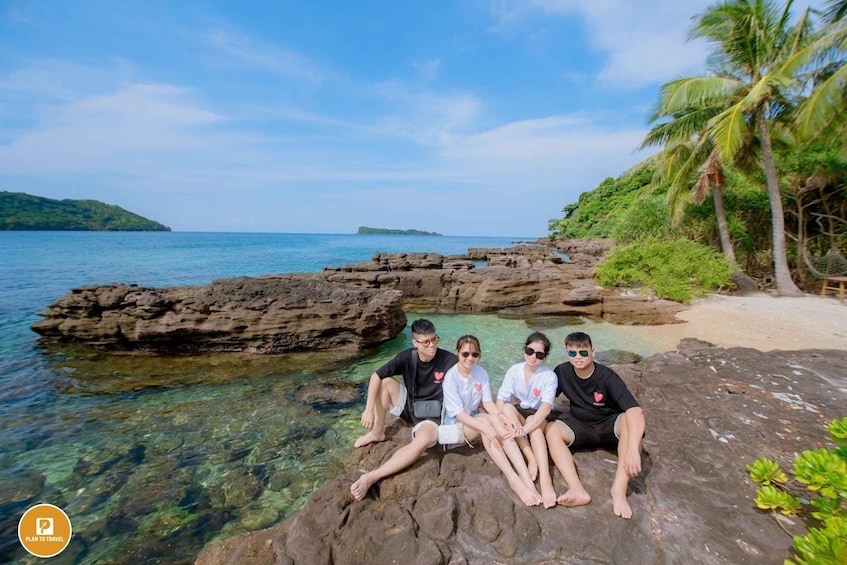 Four people sitting on a rocky outcrop by a clear blue sea with palm trees and a distant island. 4